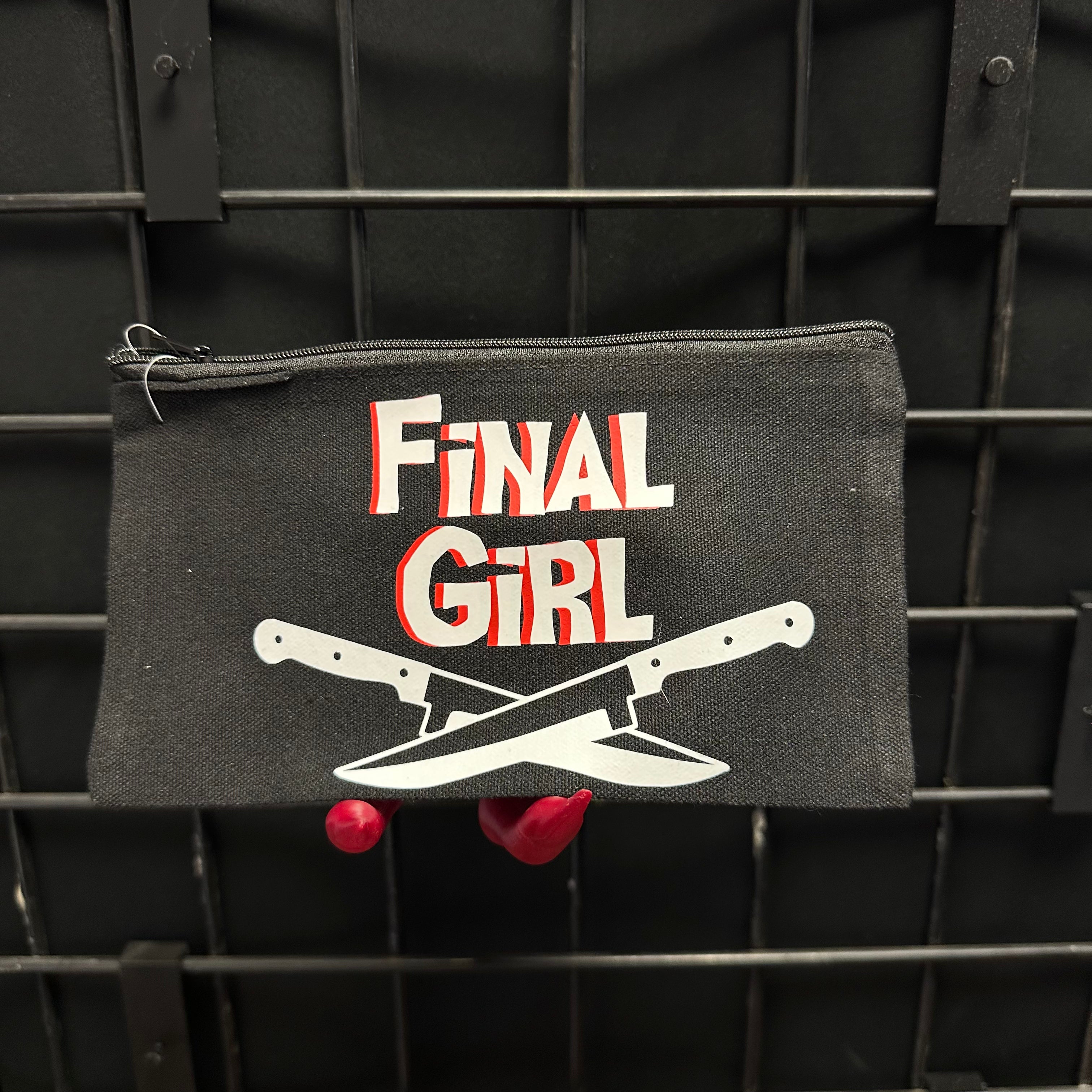 A black canvas zippered pouch featuring the 'Final Girl'  graphic with white and red text and crossed knives, displayed on a black wire rack.
