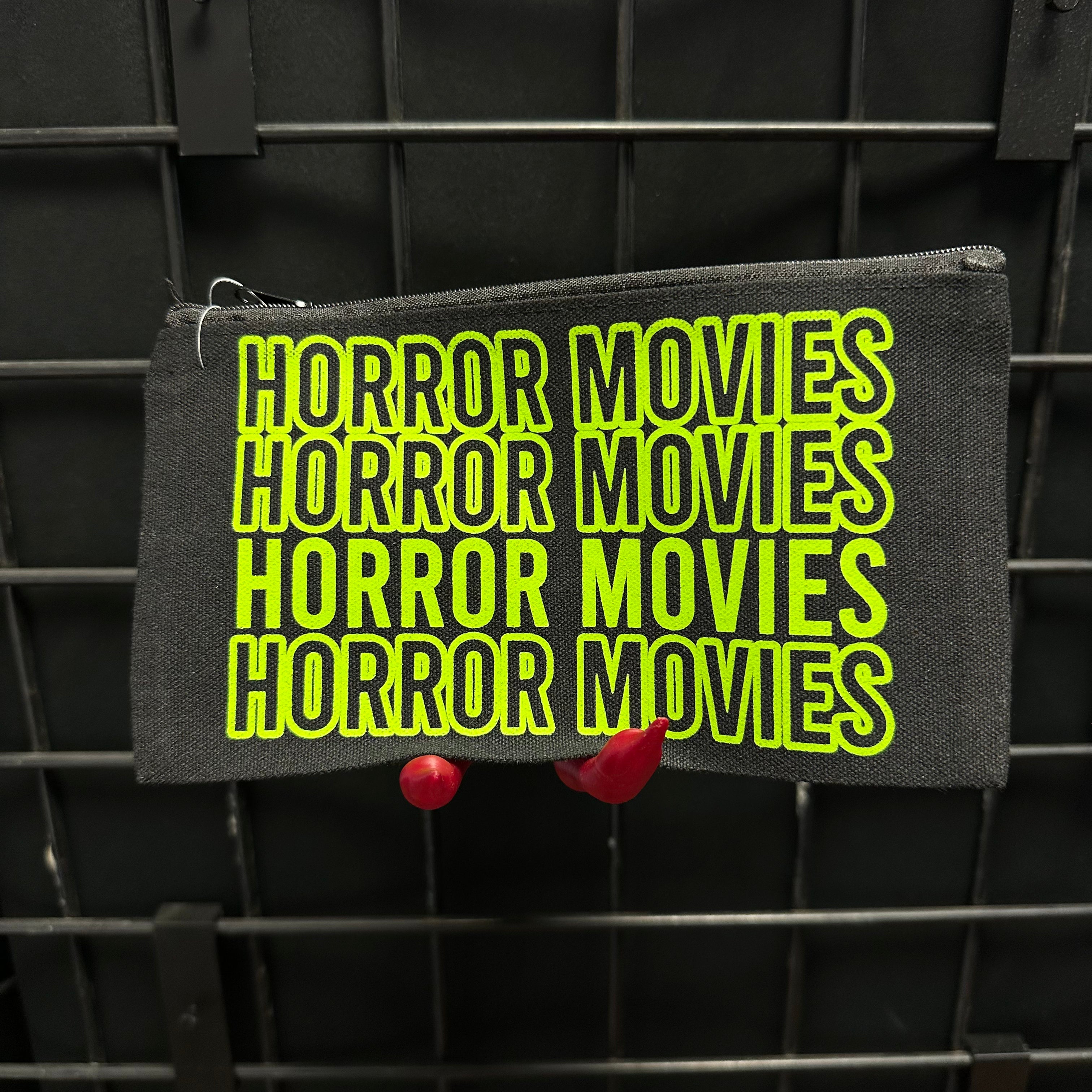 A black fabric pouch with 'HORROR MOVIES' printed repeatedly in bright neon yellow-green, hanging against a black wire rack held by one of our monster hands