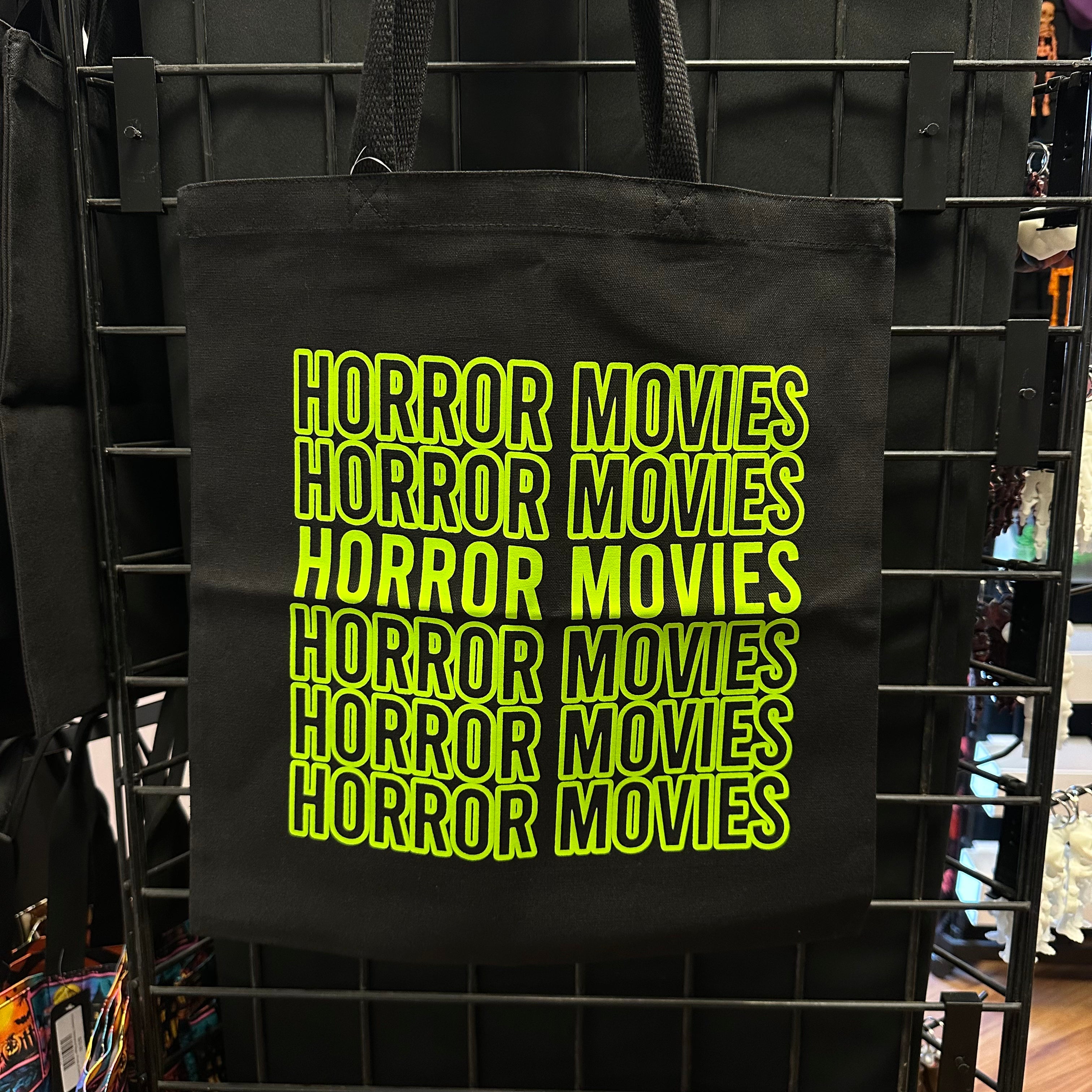 A black canvas tote bag hanging on a black metal retail display rack, featuring the phrase 'HORROR MOVIES' repeated in a bright neon yellow-green outline font.
