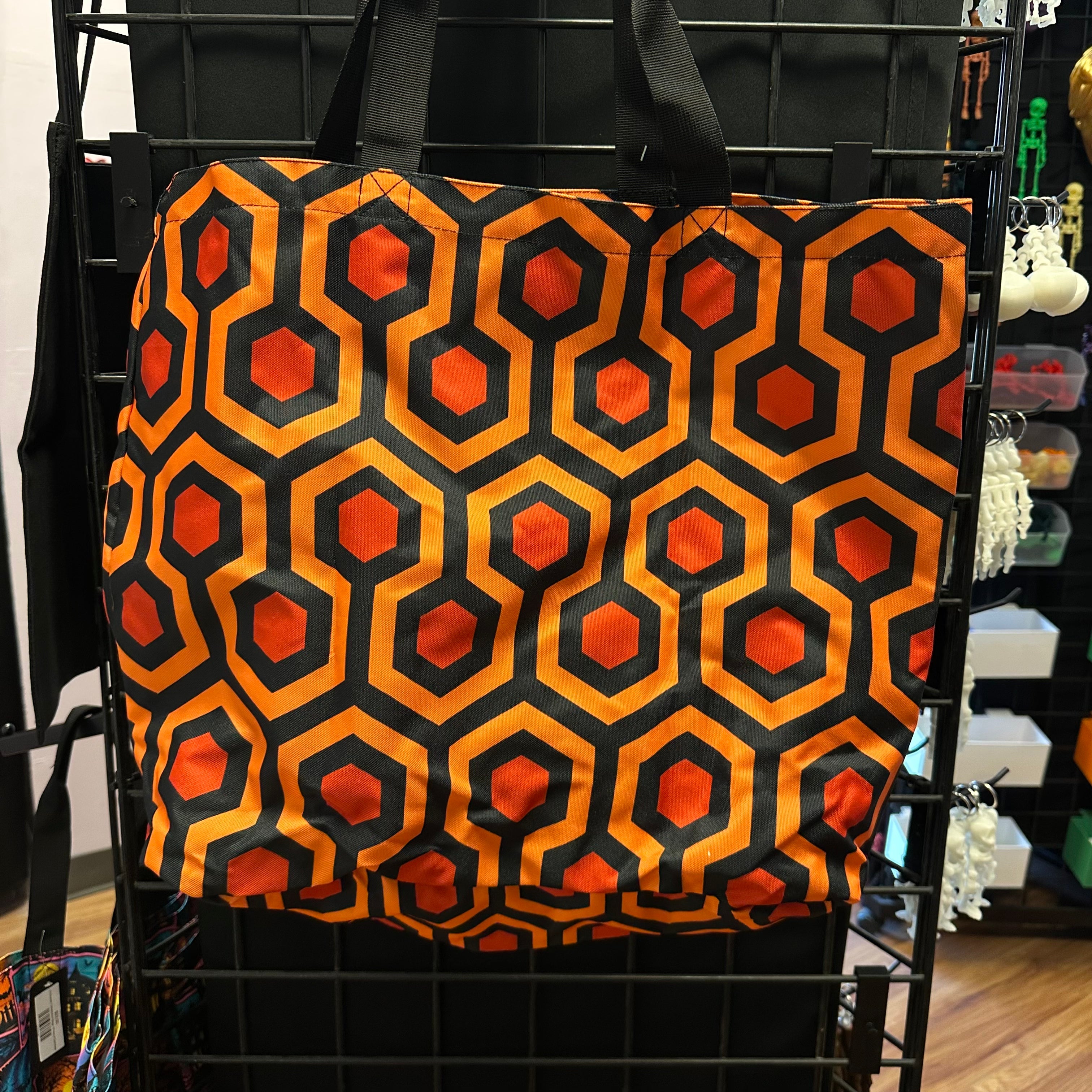 A tote bag with the overlook geometric pattern of orange and black hexagons, hanging on a black wire display rack.