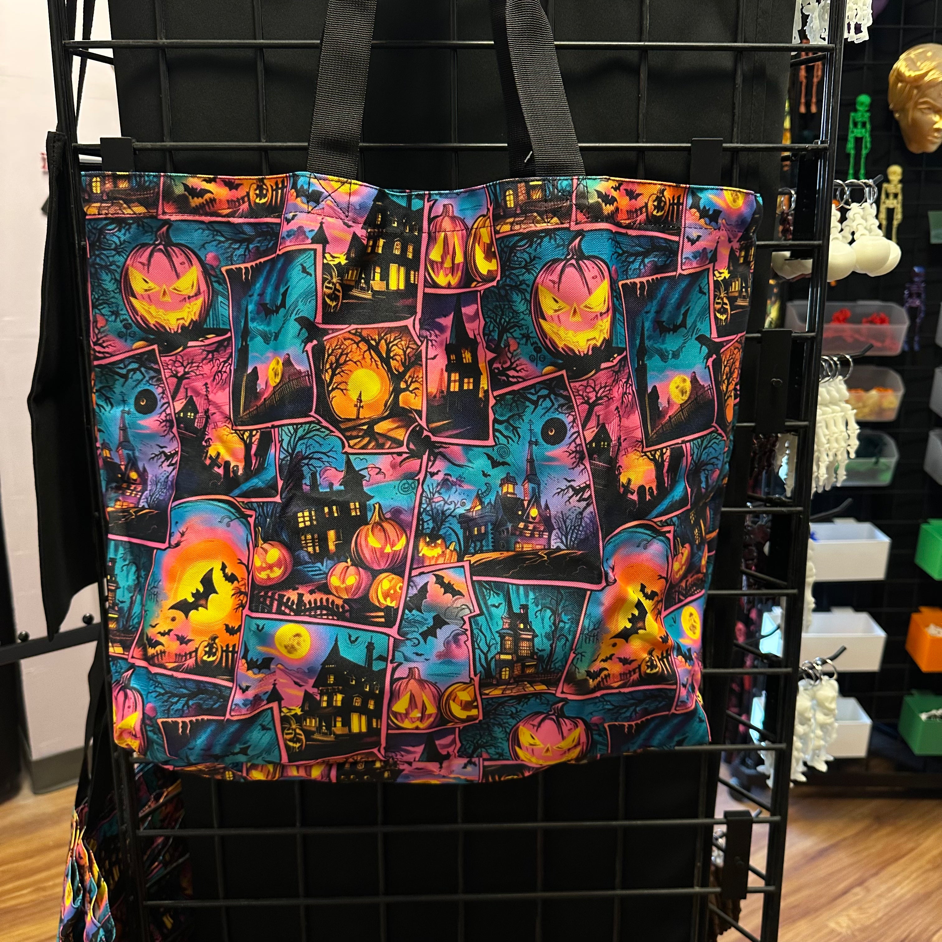 A black tote bag with a vibrant Halloween collage print featuring jack-o'-lanterns, haunted houses, bats, and pumpkins in pink, orange, yellow, and blue, hanging on a black wire display rack.